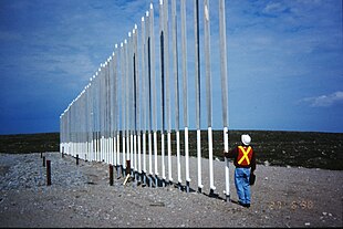 A worker in high visibility clothing and a hard hat examines a long line of pipes about four times his height sticking out of rocky ground.