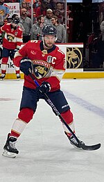 An ice hockey player skates on the ice in warmups. He has a red jersey and blue/black equipment.