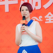 Hai Qing wearing a white sleeveless top with a bright blue collar, and a matching blue skirt, standing onstage, holding a microphone in front of her