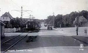 A person stands at the corner of where two roads meet, observing a trolley passing in front of houses, while some automobiles approach the intersection.