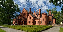 Seitliche Farbfotografie von einem braunen Schloss mit mehreren Dachgiebeln und kleinen Türmen. Rechts ist ein runder Eckbau. Die meisten Fenster haben schmale Rundbögen und der Eingang ist überdacht. Um das Gebäude sind Grünfläche und Bäume.