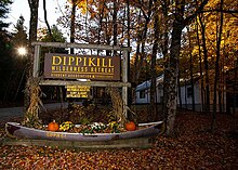 A wooden sign prominently features the retreat's name and identifies it as private property, accessible only to SUNY Albany affiliates. Below the sign, there is a canoe decorated with cornstalks and pumpkins, suggesting the photo was taken in the fall. The surrounding area is wooded with autumnal foliage, creating a rustic and seasonal atmosphere. The retreat appears to be a secluded and tranquil location for students and staff.