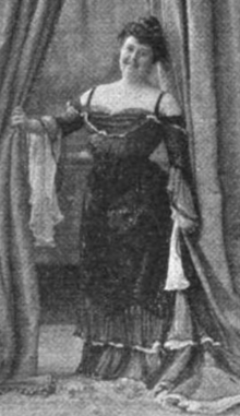 A smiling woman with fair skin and dark hair in an updo, wearing a dark gown with bare shoulders; She is standing between curtain panels, possibly on a stage