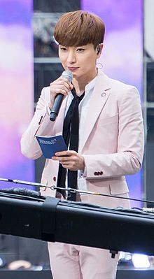 Leeteuk holding cue cards in his left hand and a microphone in his right hand close to his face, looking slightly downward