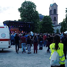 Photo von einer Person die eine gelbe Warnweste mit der Rückenaufschrift „Ordner“ trägt in der Nähe einer Konzertbühne.