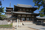 Large two-storied gate with a hip-and-gable roof and a railed open veranda on the upper floor. There are two guardina statues on either side of the passage.