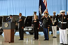 Obama and Clinton at a somber occasion, honoring the Benghazi attack victims at the Transfer of Remains Ceremony, held at Andrews Air Force Base on September 14, 2012. Soldiers are standing behind Obama and Clinton, and everyone is standing on a large wooden floor with their left hands to their side and their right hands on their upper chests.