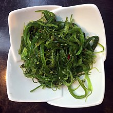 Auf einem weißen Teller ist ein Salat aus knackigen grünen Algenstreifen, Sesam und kleingeschnittenem Chili angerichtet.