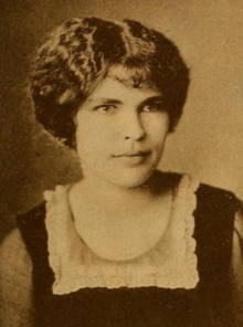 A young woman with fair skin, dark eyes, and dark wavy hair, wearing a dark dress or top with a square ruffle at the neckline