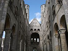 Interior shot of roofless ruined building, shot upwards towards the missing roof. The walls are in three layers, the bottom layer is made up of an arched colonnade, the second is pierced with small arched openings, and the third is mostly solid.