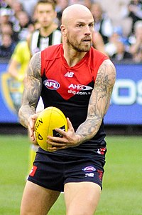 A bald, tattooed man holding a yellow football on a grassed field