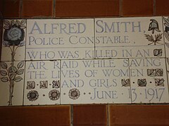 A tablet formed of five tiles of varying sizes, elaborately decorated with flowers and a stylised English policeman's helmet and bordered by yellow and blue flowers in an art nouveau style. The tablet reads "Alfred Smith, Police Constable, who was killed in an air raid while saving the lives of women and girls June 13, 1917".