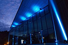 Nachtaufnahme eines modernen Gebäudes (der Tauberphilahrmonie Weikersheim) mit hoher Glasfront, die durch mehrere Flutlichtstrahler von unten blau beleuchtet wird. Das Foto ist aus der diagonalen Froschperspektive aufgenommen.