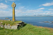 A stone cross in the Celtic style sits in a grassy field on an overgrown stone plinth. Small rocky reefs lie in the sea beyond, and there are high green hills in the distance.