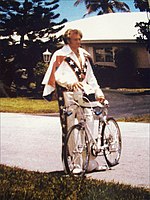 Man in his forties, standing in front of a house and holding a bicycle and helmet. He is smiling and wearing a red, white, and blue jumpsuit.