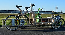 Lastenrad mit hölzernem Rahmen, dahinter ein weiteres. im Hintergrund ein Flughafengebäude