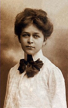 A young white woman with hair in a bouffant updo, wearing a white pleated blouse with a dark bow at the neck