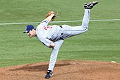 A man in a gray baseball uniform and navy-blue cap follows through after throwing a baseball. He is standing with his right leg planted on a dirt mound within a grass field and his left leg raised in the air behind him.