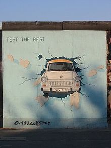 Frontale Farbfotografie von der Berliner Mauer mit einem Bild von einem weißen Auto, dem Trabant, der eine hellblaue Wand in Richtung Betrachter durchbricht. Links oben steht „Test the best“.