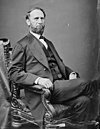 A bearded man in a dark formal suit sits on an ornate wooden chair