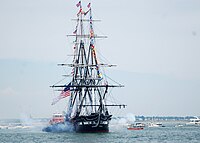 A photo of a ship without sails. There is white cannon smoke emitting from the left and right sides of the ship. A tugboat is alongside