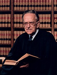 An older man with glasses wearing black with a shelf full of brown, red and black books behind him