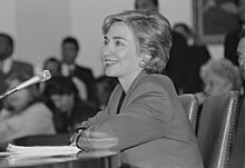 Photograph of Clinton making a presentation sitting at a table in front of a microphone