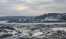 An aerial view of a small settlement beside a wide river, covered in a layer of snow. A large forested hill lies on the other side of the river.