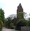 A tall narrow tower with a small pointed spire surrounded by trees in leaf almost hiding the body of the church; to the left is a footpath with railings and a wall