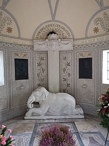 Heller, runder Innenraum mit einem christlichen Kreuz unter dem ein Elefant liegt. Auf dem Kreuz ist ein Jesushaupt mit Dornenkrone zu sehen, darüber der Spruch "Ich bin der Anfang und das Ende"