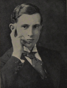 A clean-shaven young man in a suit holding his hand loosely on the side of his head