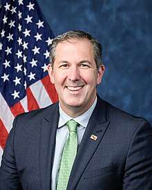 Official House portrait of Olszewski smiling in front of the U.S. flag, wearing a black suit with Maryland flag lapel pin, light blue shirt, and green tie.