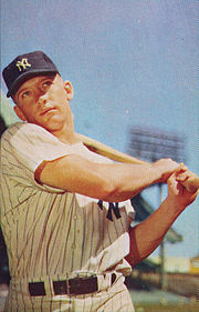 "A young man in New York Yankees pinstripes and cap looks ahead whilst swinging a baseball bat."