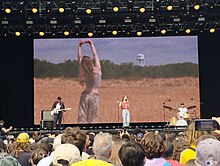 Cain and a band performing on a stage in front of a screen showing visuals of herself in a field