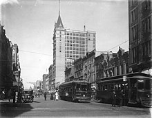 A black-and-white photograph of old-style streetcars traveling in the center of a street lined with multi-story buildings. People can be seen boarding the streetcars.