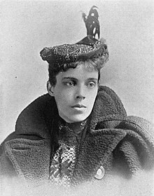 A young white woman wearing a flat cap with a plume, and a bulky coat with wide lapels.