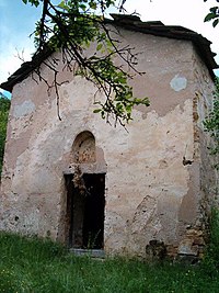 The west wall of a basically-constructed stone medieval church with a gabled roof. A door with a semicircular niche on top of it is visible on the wall.