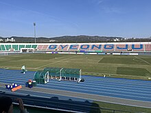 A football pitch surrounded by an athletics track within a small stadium with tiered seating all around. Among mostly orange seats, blue seats spell out the city name 'Gyeongju'