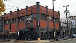 Iso image of the beer hall showing the west and north faces. This is a three story building. The bottom floor is painted black and has street facing windows. The second and third floor are exposed red brick, with multi-paned windows on the third floor. On the corner in front of the entrance is a sandwich board sign saying "Loyal Legion"