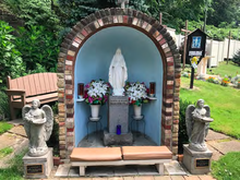 A photo of a shrine with a white figure, representing Mary, in the center of an alcove. By each side of this figure there is a vase of flowers and a statue of an angel.