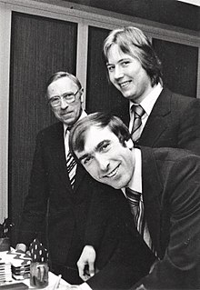 A black and white image of three white men in suits about to sit down at a small table against a wall, perhaps at a banquet. The photo is probably from the 1960s or 70s. Two are about 30 and one is about 60 years old.