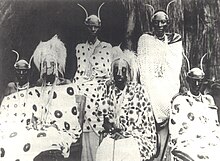 A group of Rwandan royal family members sit and stand in front of a hut, adorned in their royal regalia.