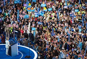A view looking downward at a raised blue stage. A woman in white pantsuit stands behind a podium (and clear safety shields) facing a crowd of supporters holding American flags, "Hillary" signs, etc.