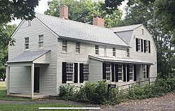 A two-story gray wooden house with light green shutters seen in winter, with snow on the ground. A long wing with two brick chimneys projects towards the left from a higher rear section. At lower right is a black mailbox with "5Ave" on it next to a clear paved driveway