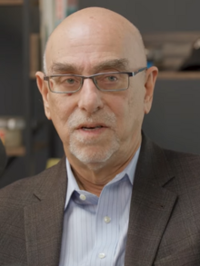 Ruy Teixeira wearing a light striped shirt and a gray suit jacket, looking left of camera and appearing to speak