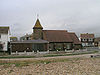 Side view of a small brick church with a steeply pitched, red-tiled roof extending to just above ground level. Below the roof, there are three paired windows separated by buttresses. A round tower with a conical spire topped by a crucifix stands at the left end. In front of this, a low, flat-roofed extension in dark brick, with a nine-light ground-to-roof window. A shingle beach runs in front.