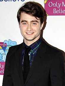 Frontale Farbfotografie von einem jungen Mann, der lächelnd vor einem Plakat steht. Seine dunklen Haare sind nach links gekämmt und er trägt einen schwarzen Anzug mit blau-grün-kariertem Hemd und schwarzer Krawatte.