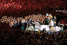 Coldplay stand on a stage surrounded by a large crowd of fans