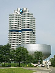 A tall building formed by cylinders stands beside a short bowl-shaped building, there are trees near both of them as well