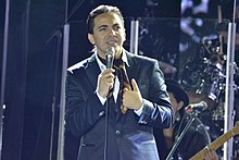 A man wearing a dark blue suit is holding a microphone with his right hand